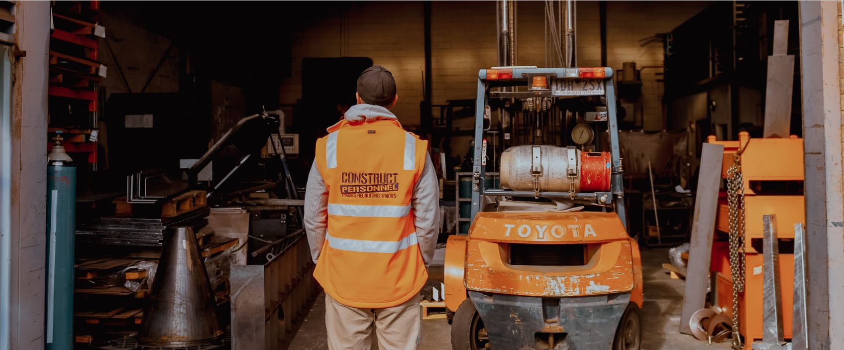 Worker in a high-visibility vest standing in a workshop, facing a forklift.