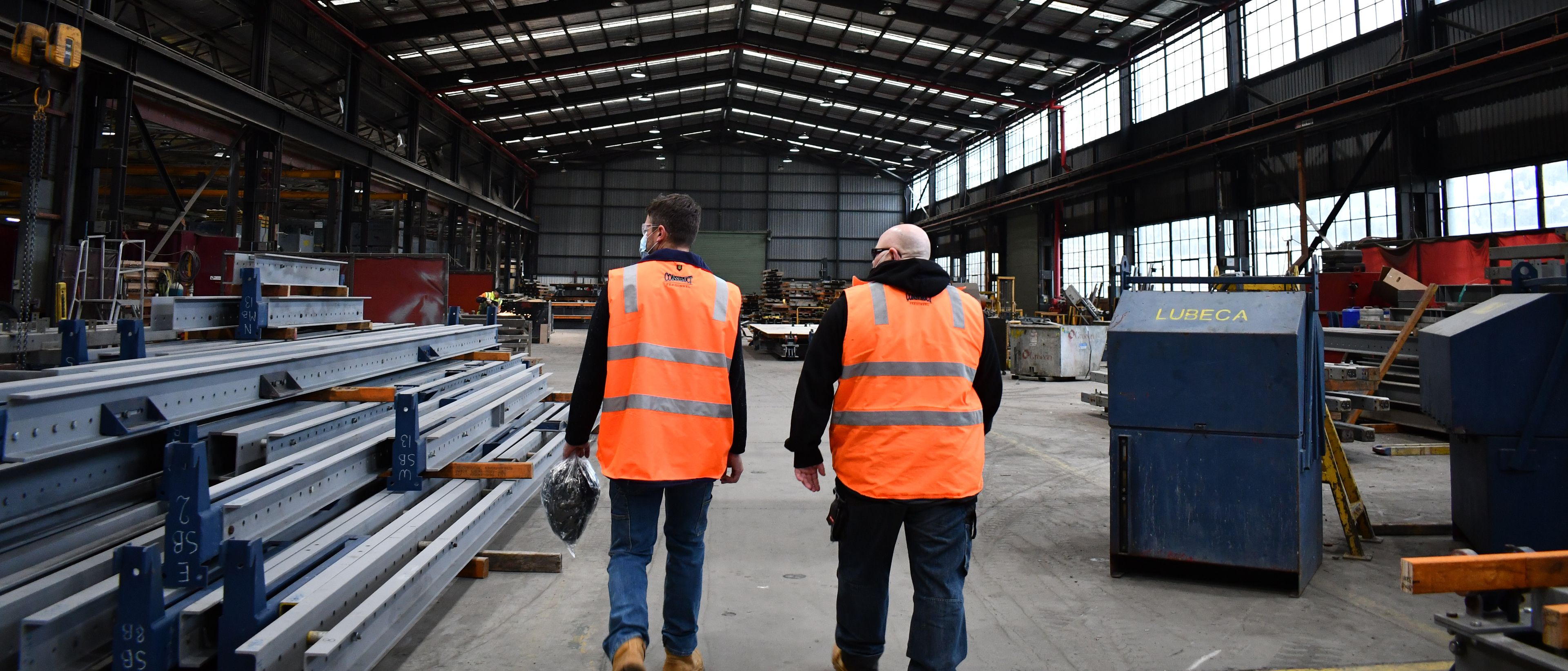 Two workers in high-visibility vests walk through a large industrial warehouse.