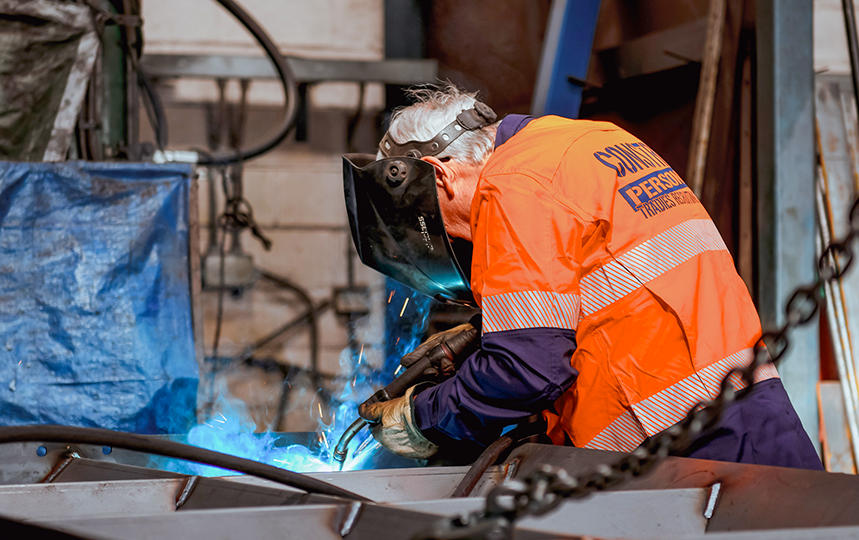 Construction worker wearing an orange high-visibility jacket and welding mask, performing welding work in a workshop